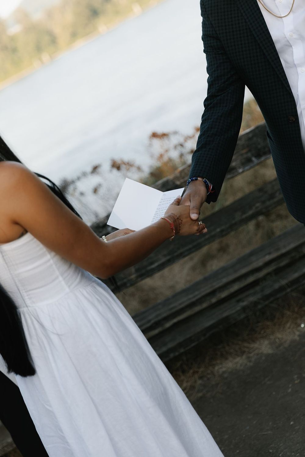 Bride in white dress holds groom's hand while reading wedding vows outdoors near a waterfront.
