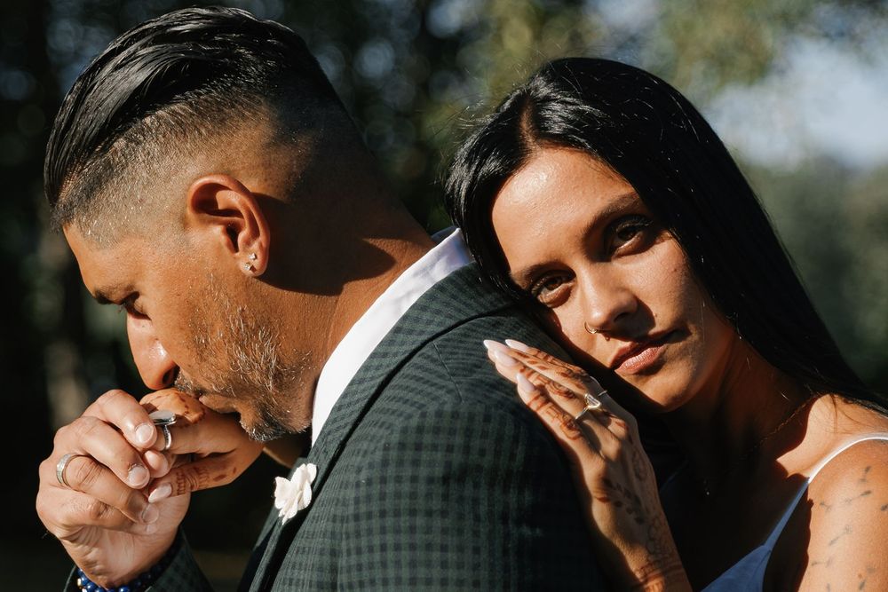 Groom kisses bride's hand as she rests her head on his shoulder outdoors.