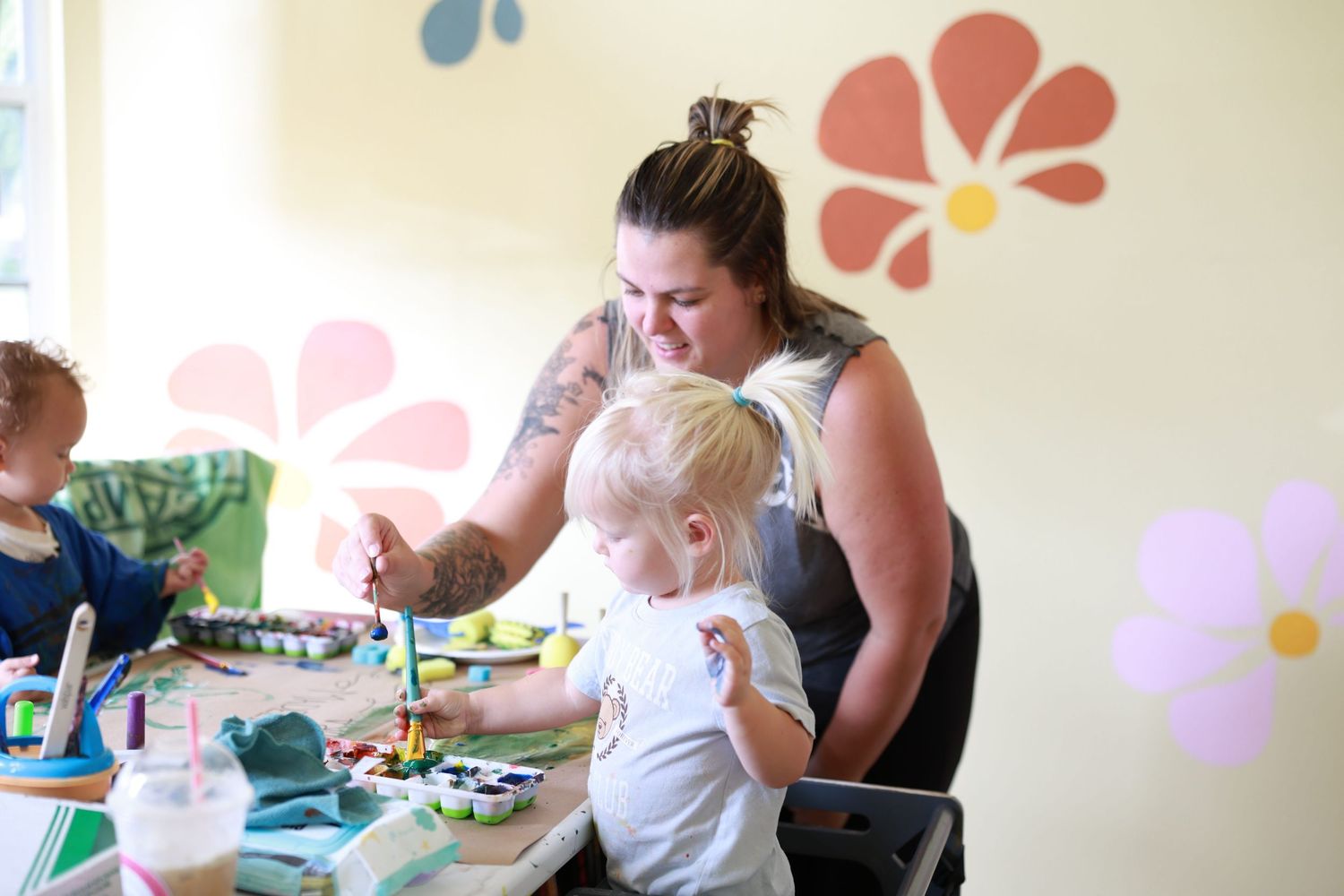 A woman with tattoos helps a toddler paint at a colorful art table with floral wall decor in a bright room. Family Photographer In Bradenton