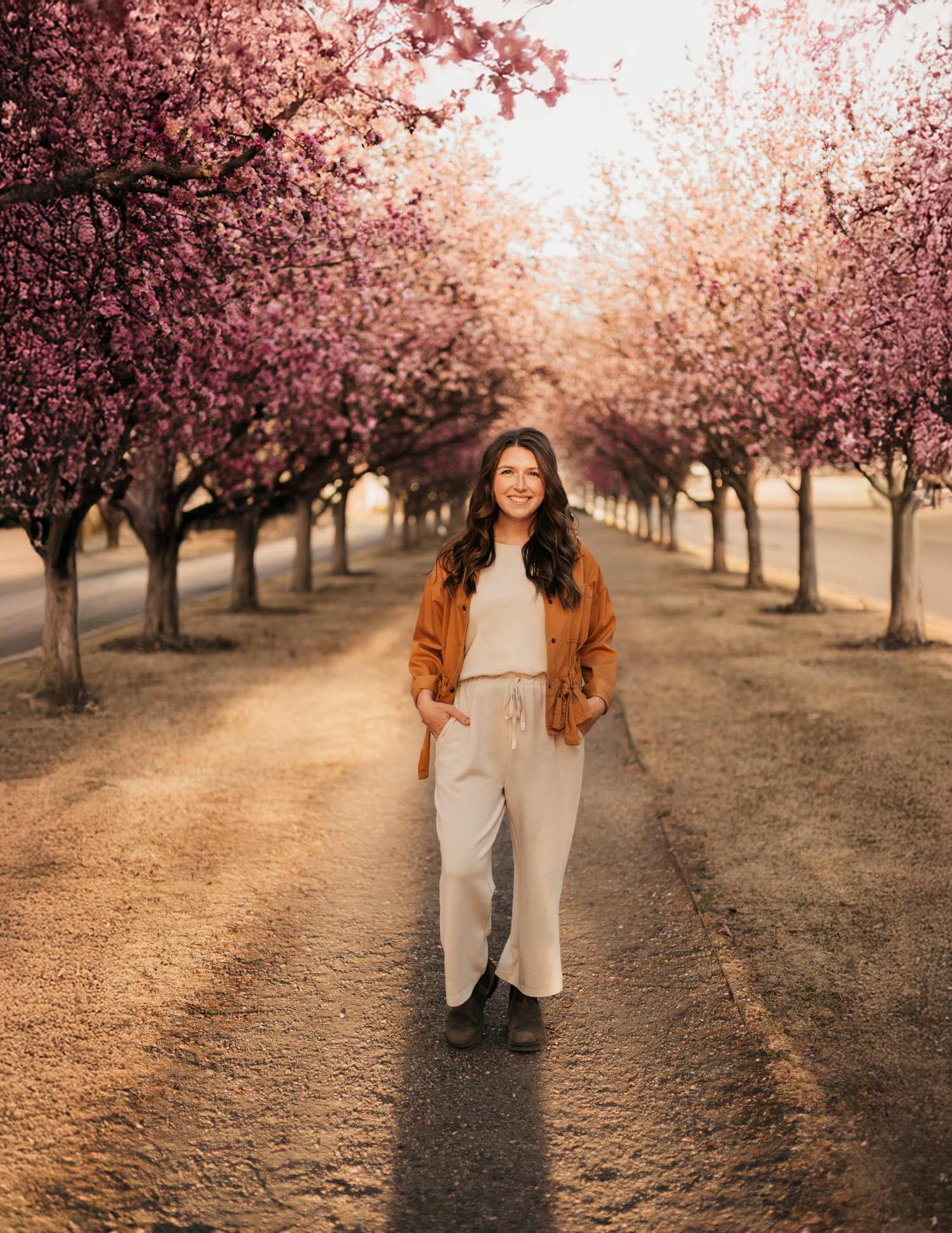 Woman smiling on a tree-lined path surrounded by blooming pink cherry blossom trees in warm golden light.
