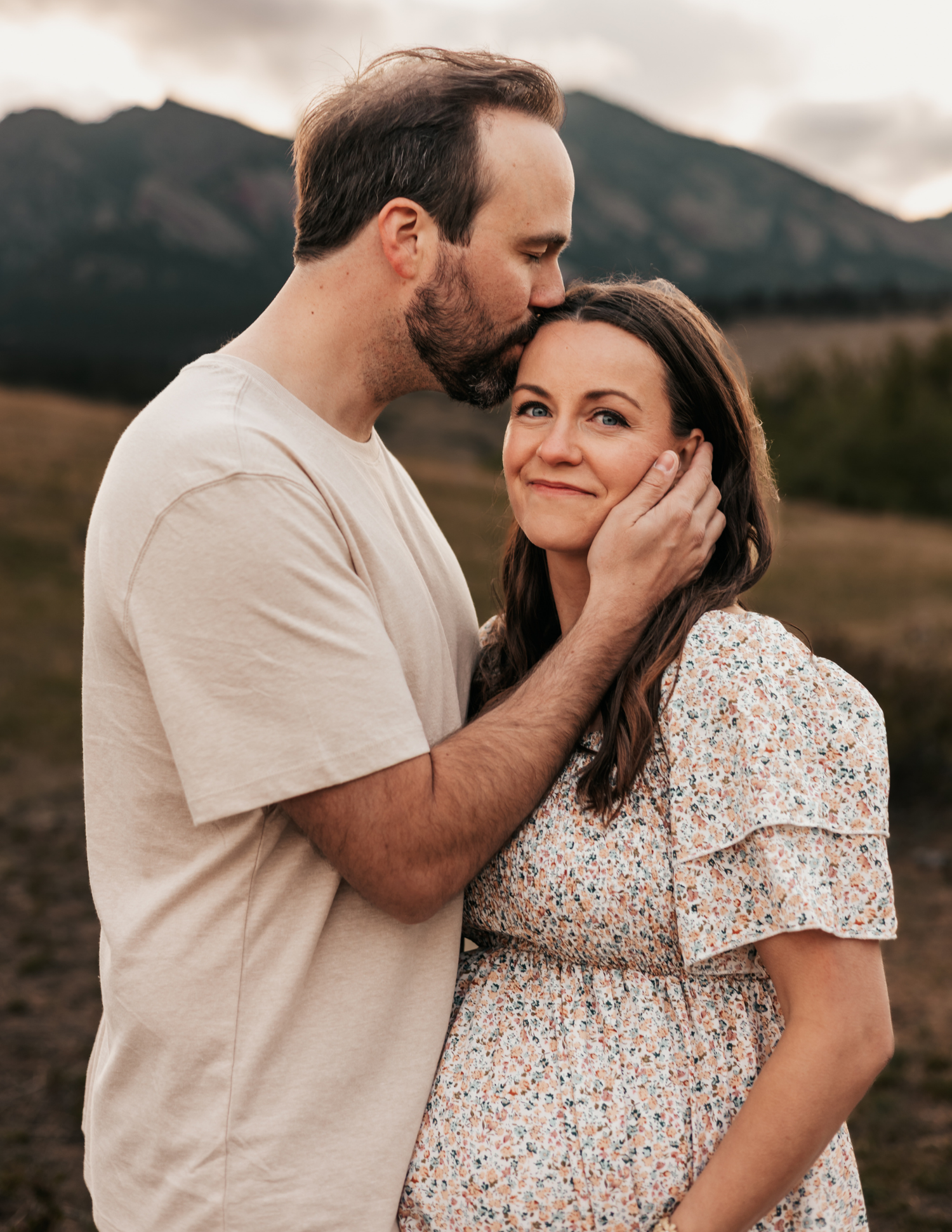 A couple shares a tender moment outdoors, man kissing woman's head with mountains in background.