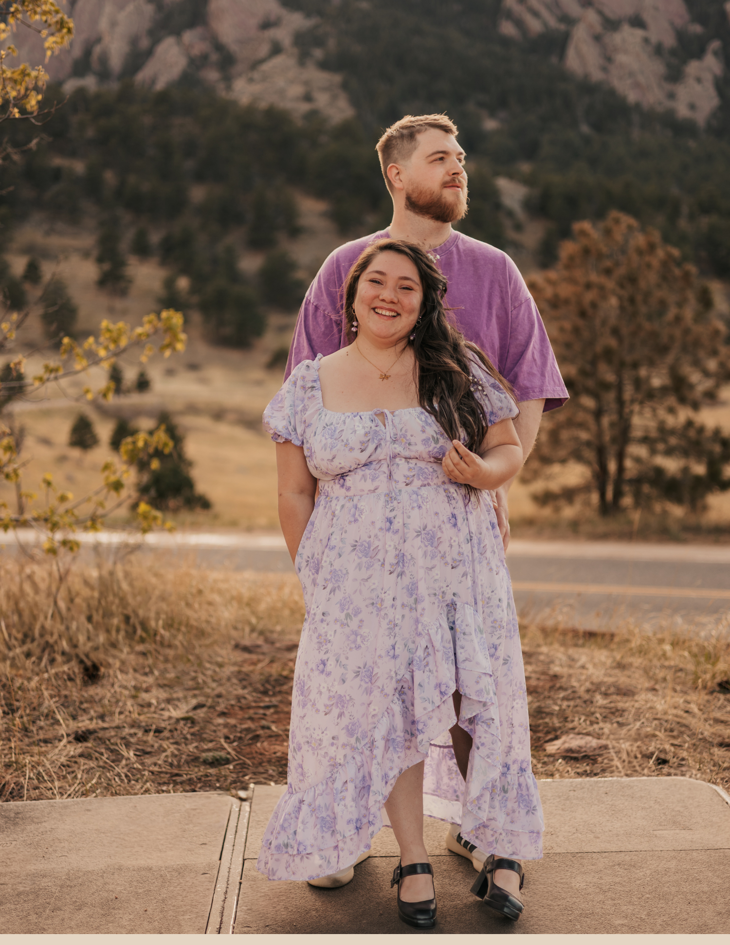 Couple posing outdoors in Colorado mountains, woman in floral lavender dress, man in purple shirt, rocky hills backdrop.