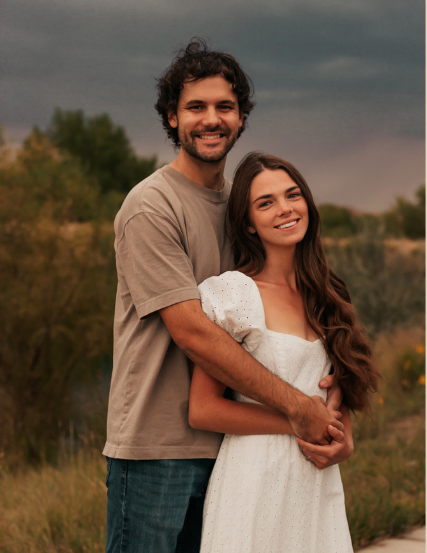 A smiling couple embraces outdoors at dusk, woman in white dress, man in tan shirt, with moody sky background.