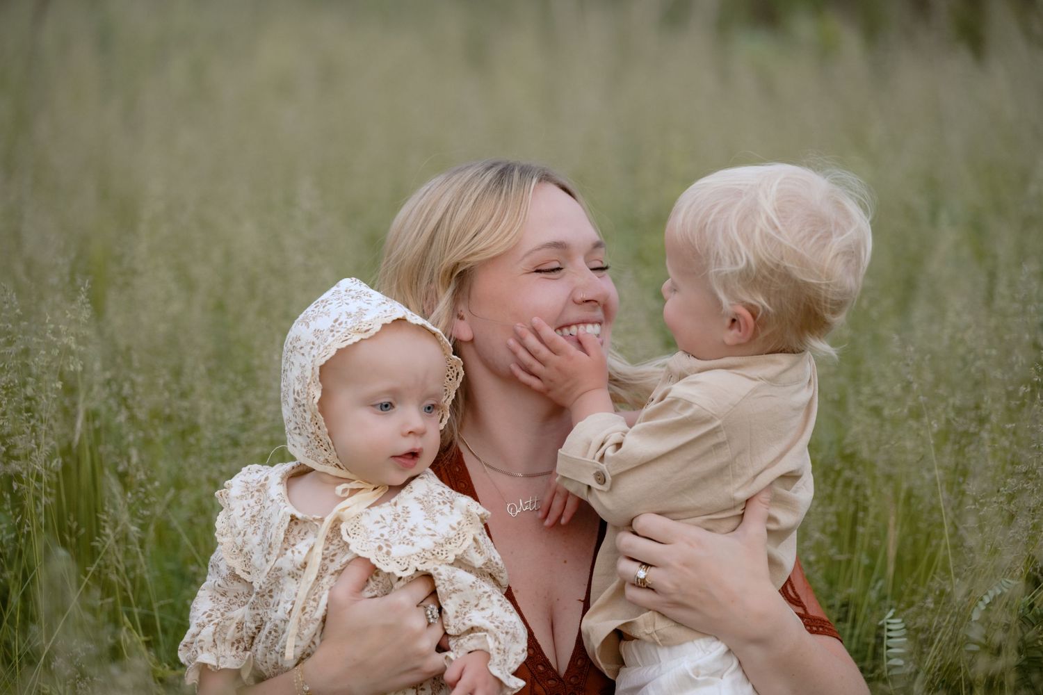 Mom sits in grassy field with her babies, toddler son holding her face and smiling during family photos in Hudson, WI.