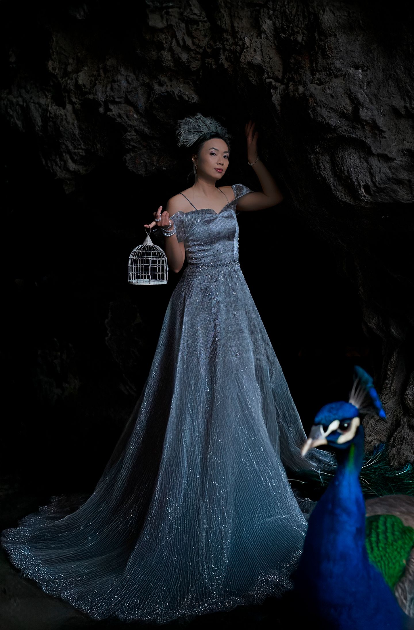 Elegant woman in silver gown holds birdcage beside peacock in dramatic cave setting.