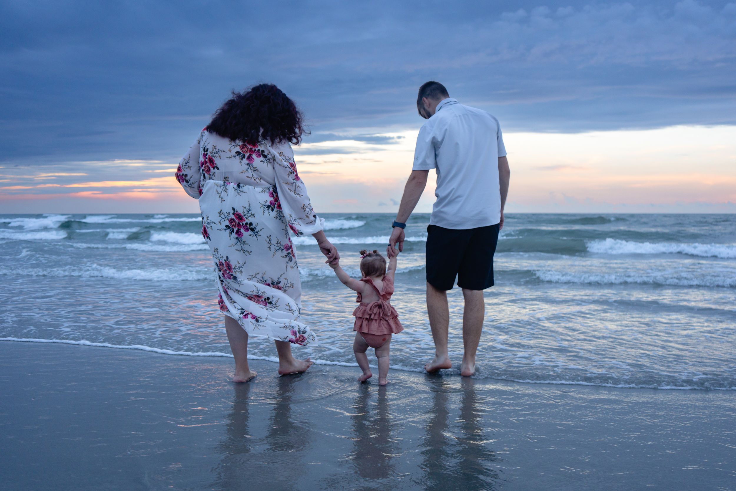 Family of three walking on beach at sunset, parents holding toddler's hands near ocean waves.