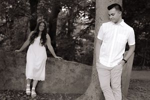Black and white photo of a couple in white clothing posing near a stone wall in a wooded outdoor setting.