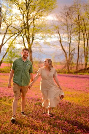A couple holding hands and walking through a field of pink wildflowers at sunset.