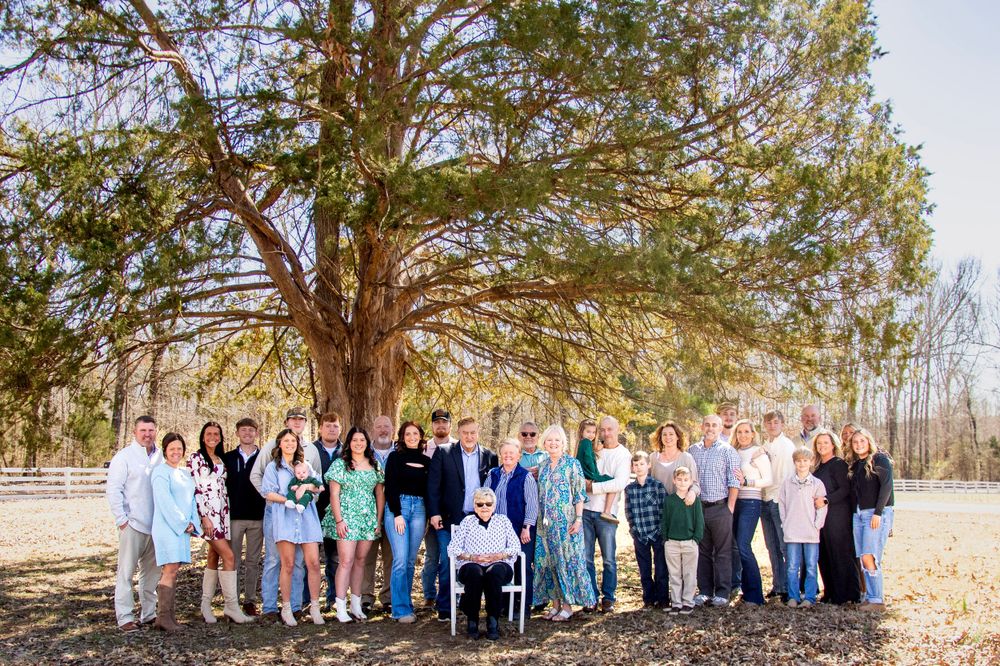 Large extended family gathered in front of a big tree during an outdoor portrait session, smiling and dressed in coordinated outfits.