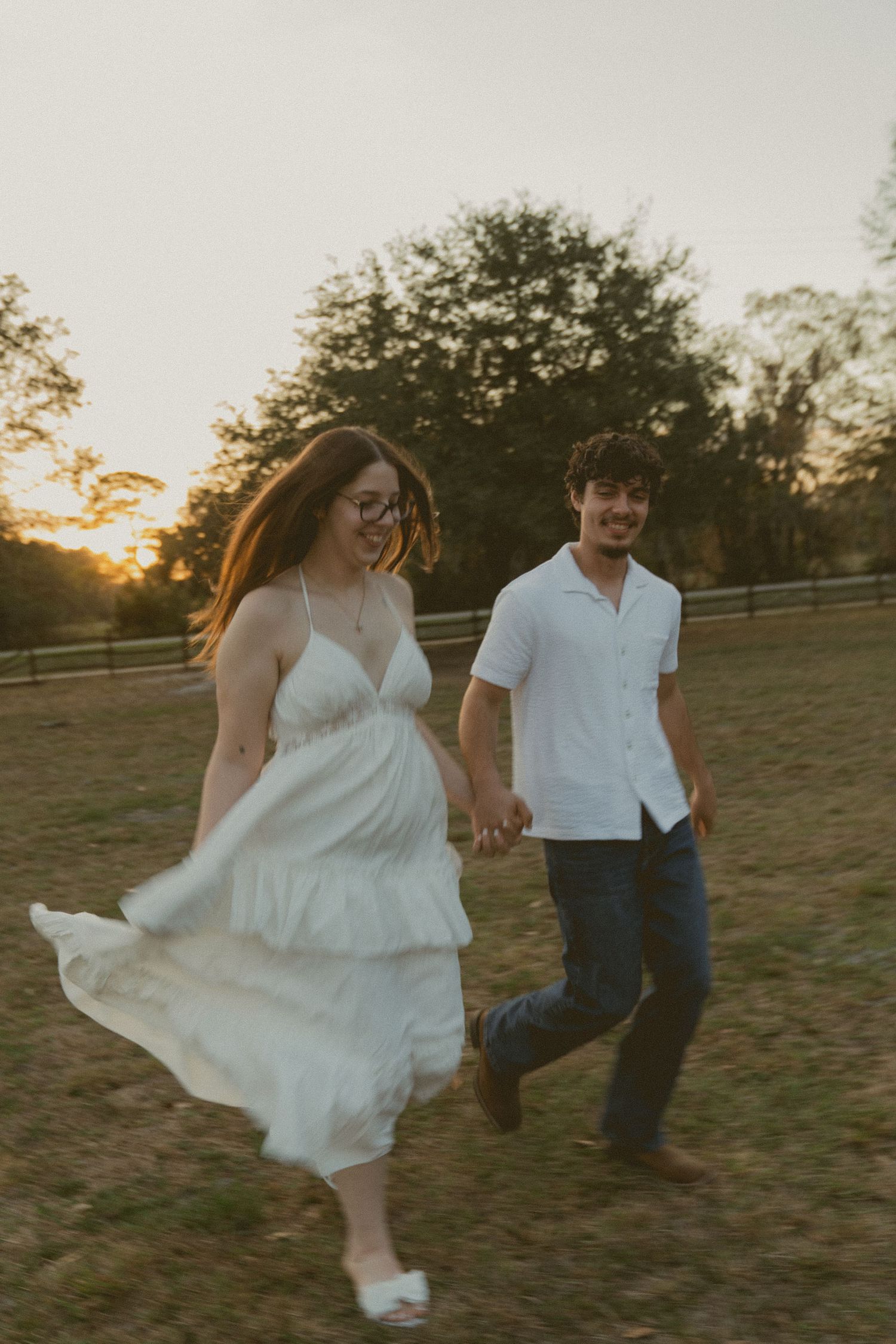 A couple in white runs playfully through a grassy field at sunset, with the bride's dress flowing in the wind.
