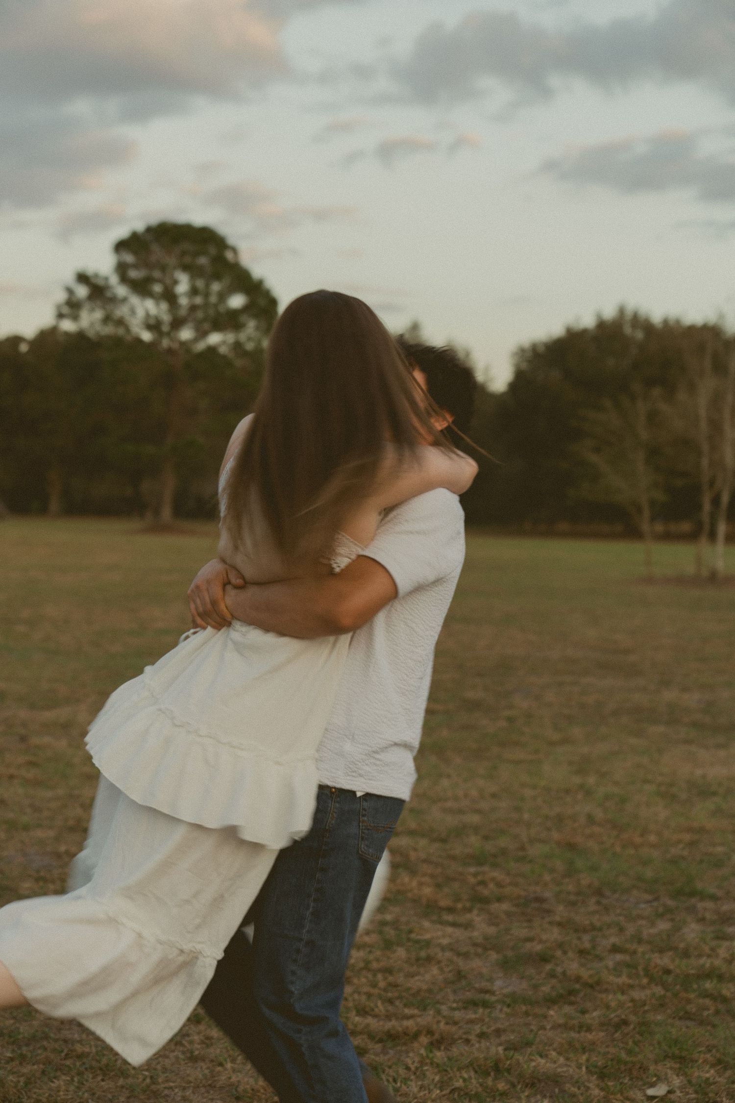 A couple in casual attire embraces and dances together in a grassy field at sunset.