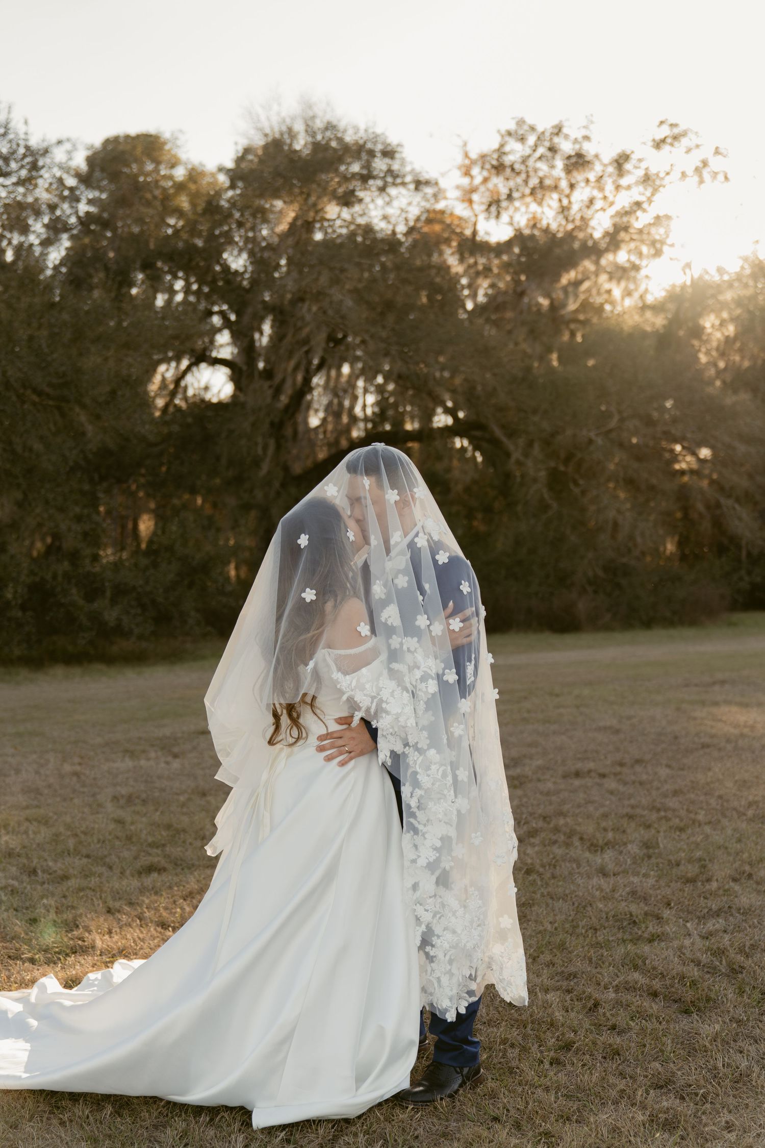 A romantic sunset wedding photo shows a couple embracing under a flowing white veil in a grassy field at golden hour.