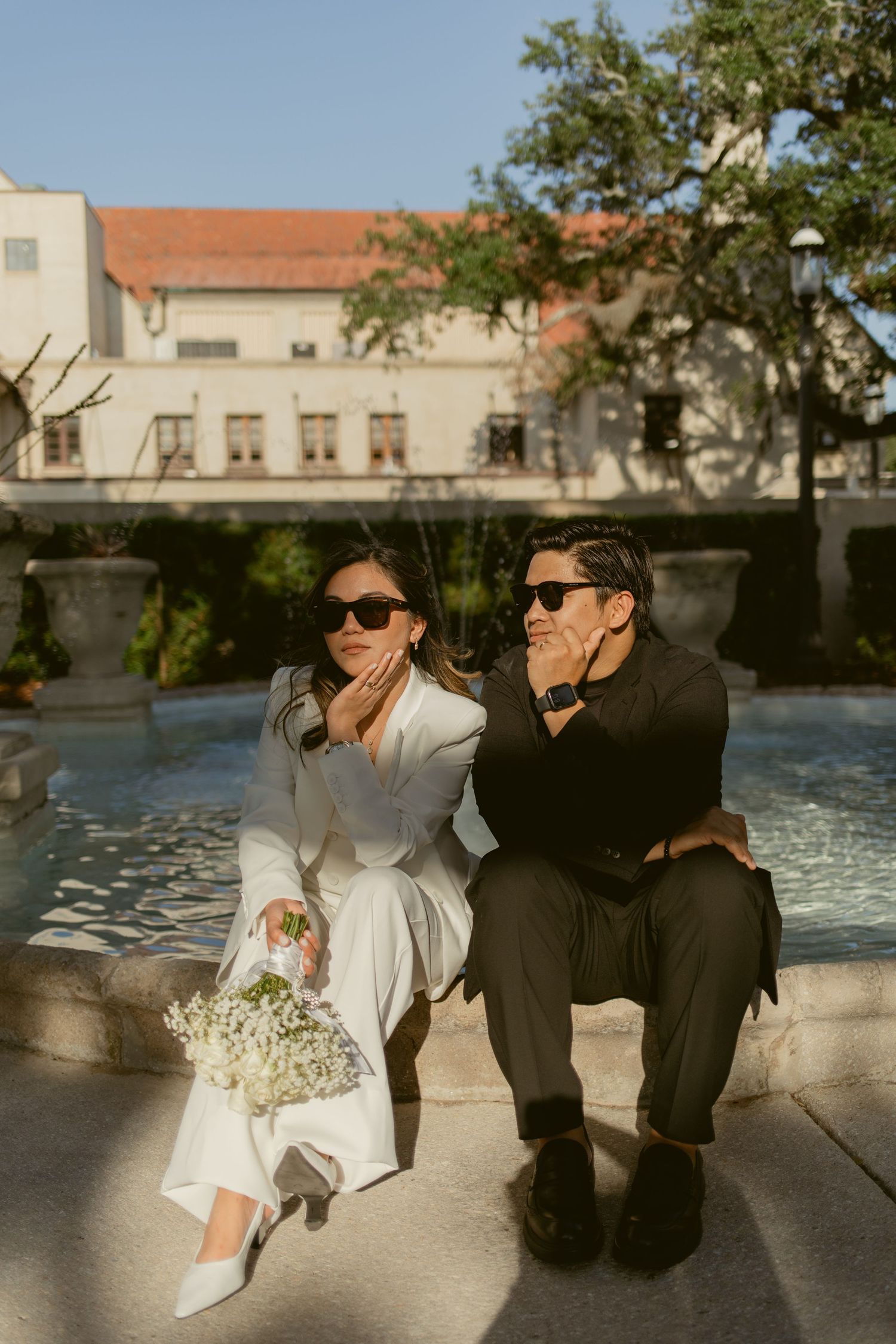 A couple in wedding attire sits playfully by a fountain on a sunny day at a historic venue.
