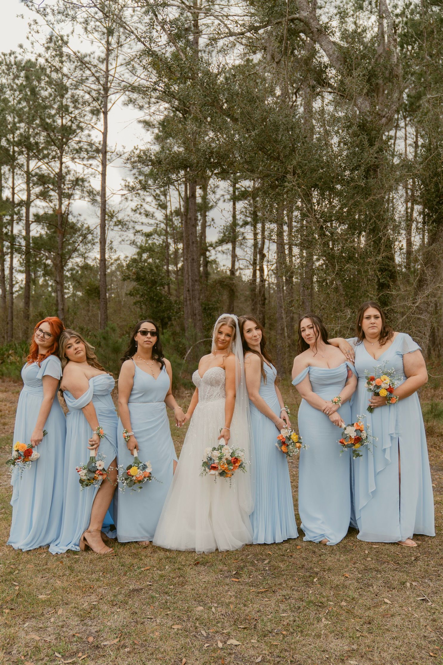 Wedding party in light blue bridesmaid dresses and white bridal gown pose together on grassy hill with trees in background.