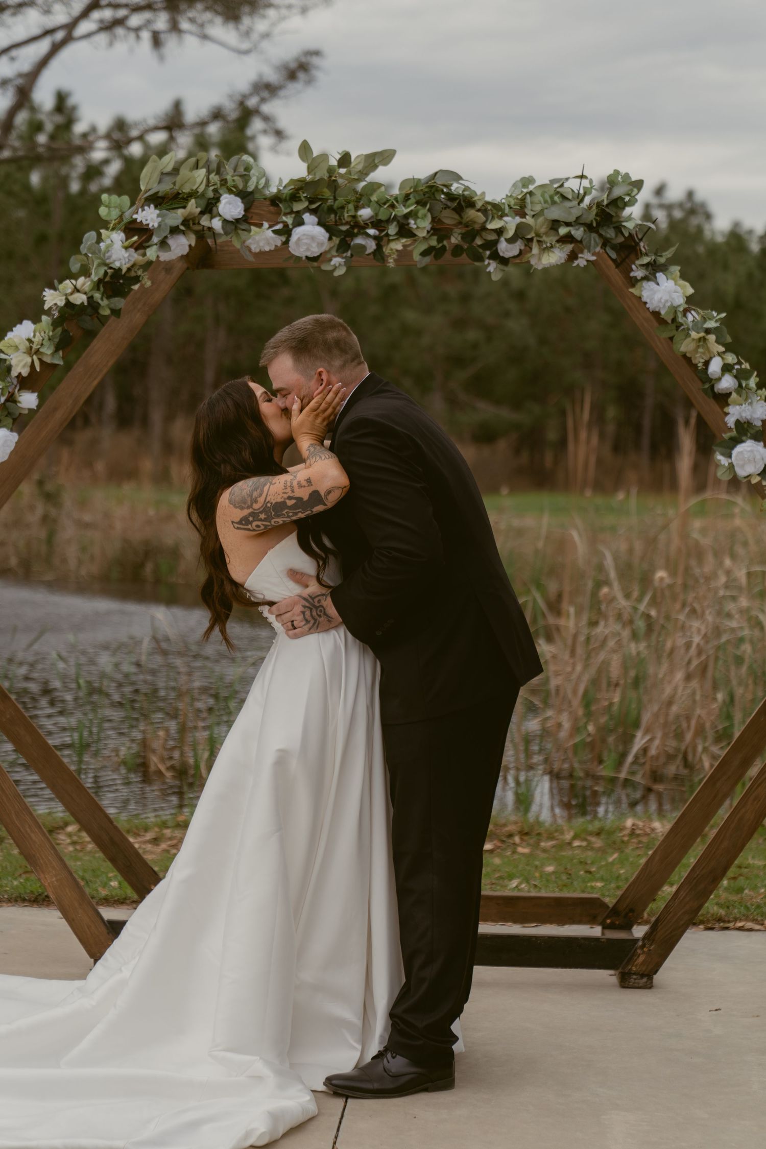 A romantic wedding couple shares an intimate kiss on a wooden deck surrounded by tall grass and natural scenery.