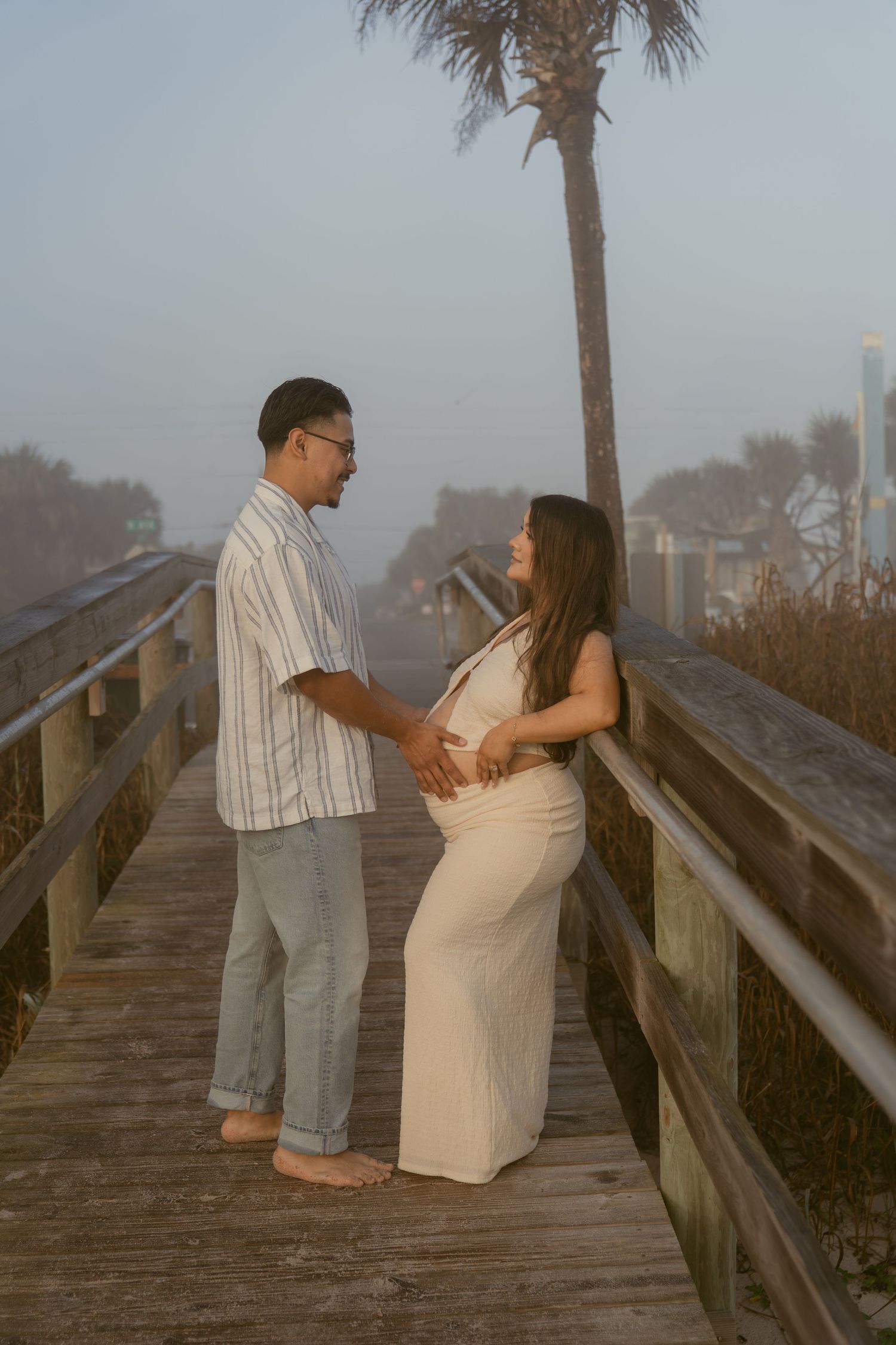 Two people share a romantic moment on a wooden boardwalk near a palm tree at dusk.