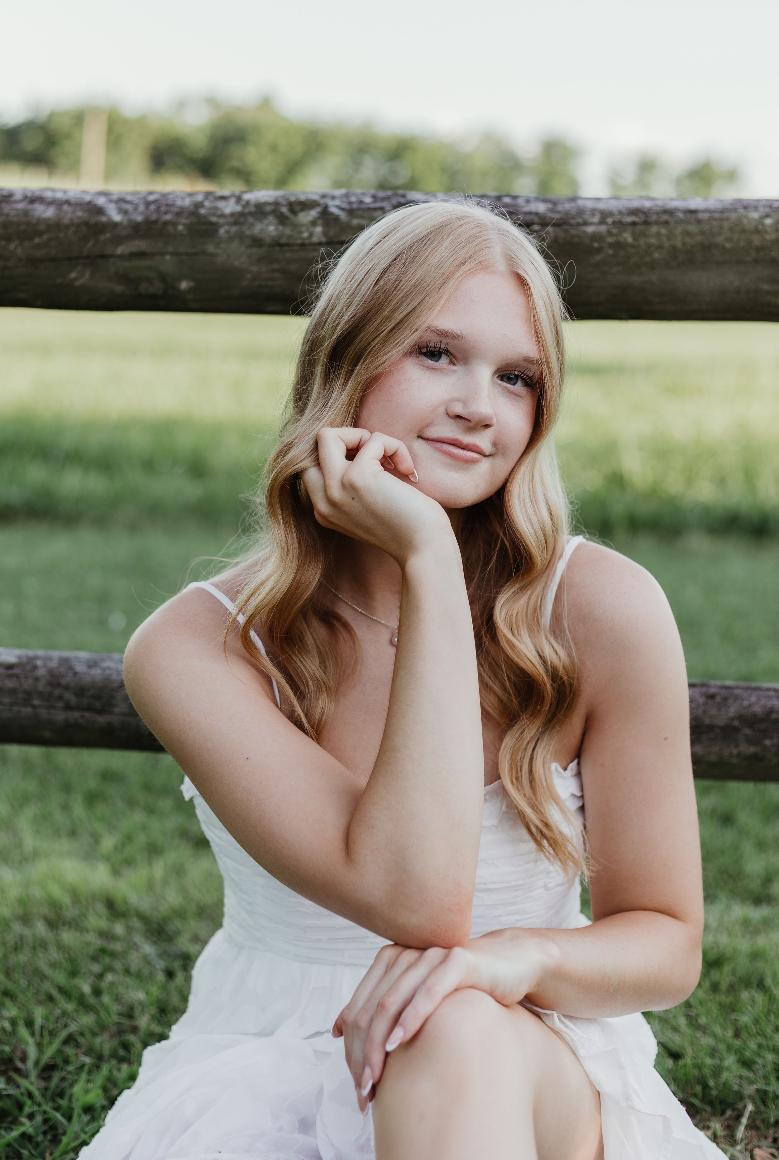 Senior in white dress sitting by rustic wooden fence in sunny outdoor setting with long wavy blonde hair.