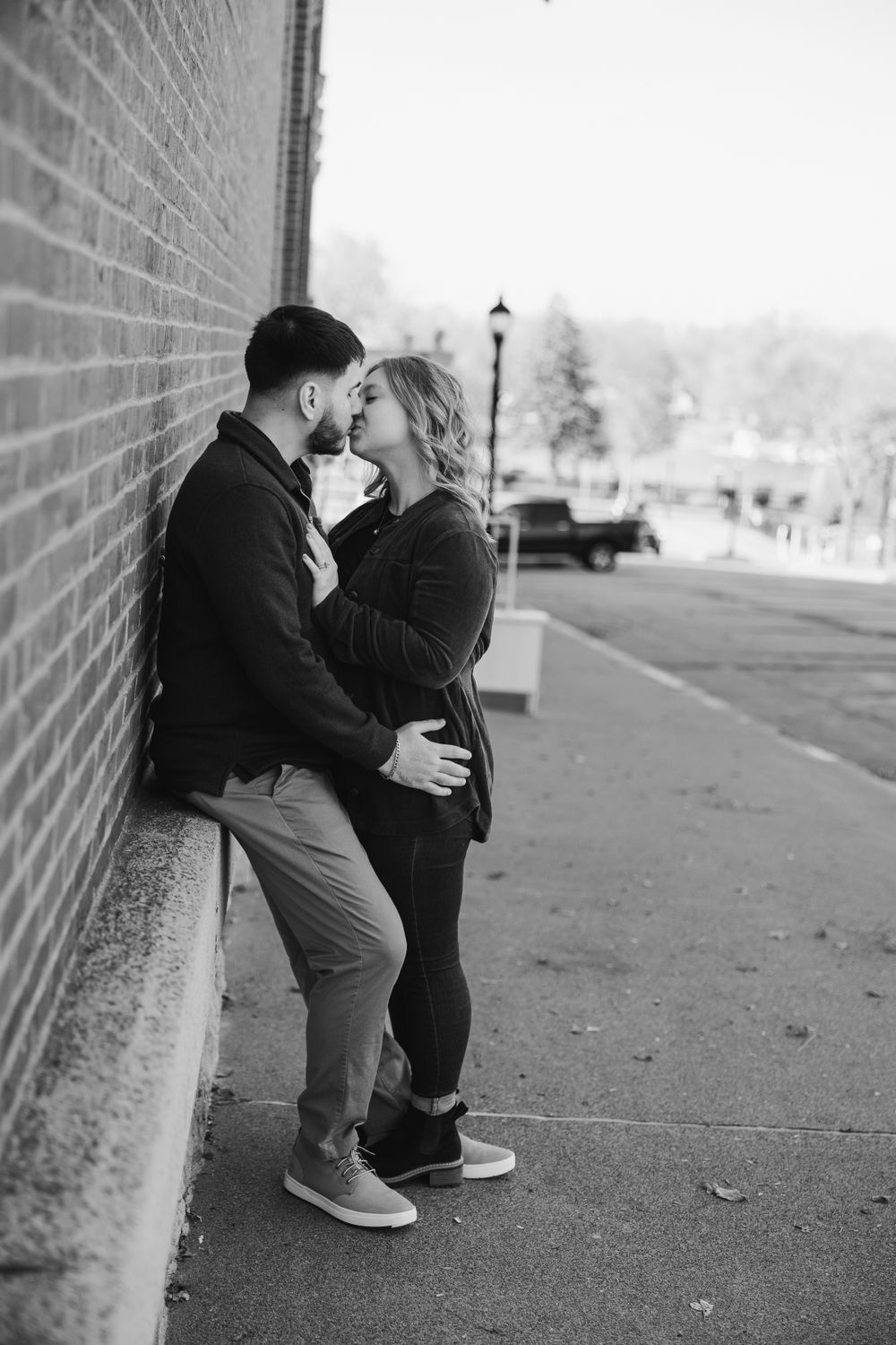 A romantic couple shares an intimate moment against a brick wall on a quiet street.