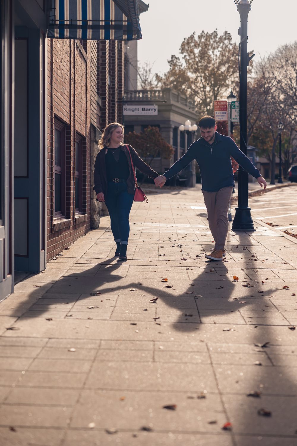 A couple walks hand in hand down a sunlit sidewalk casting long shadows.