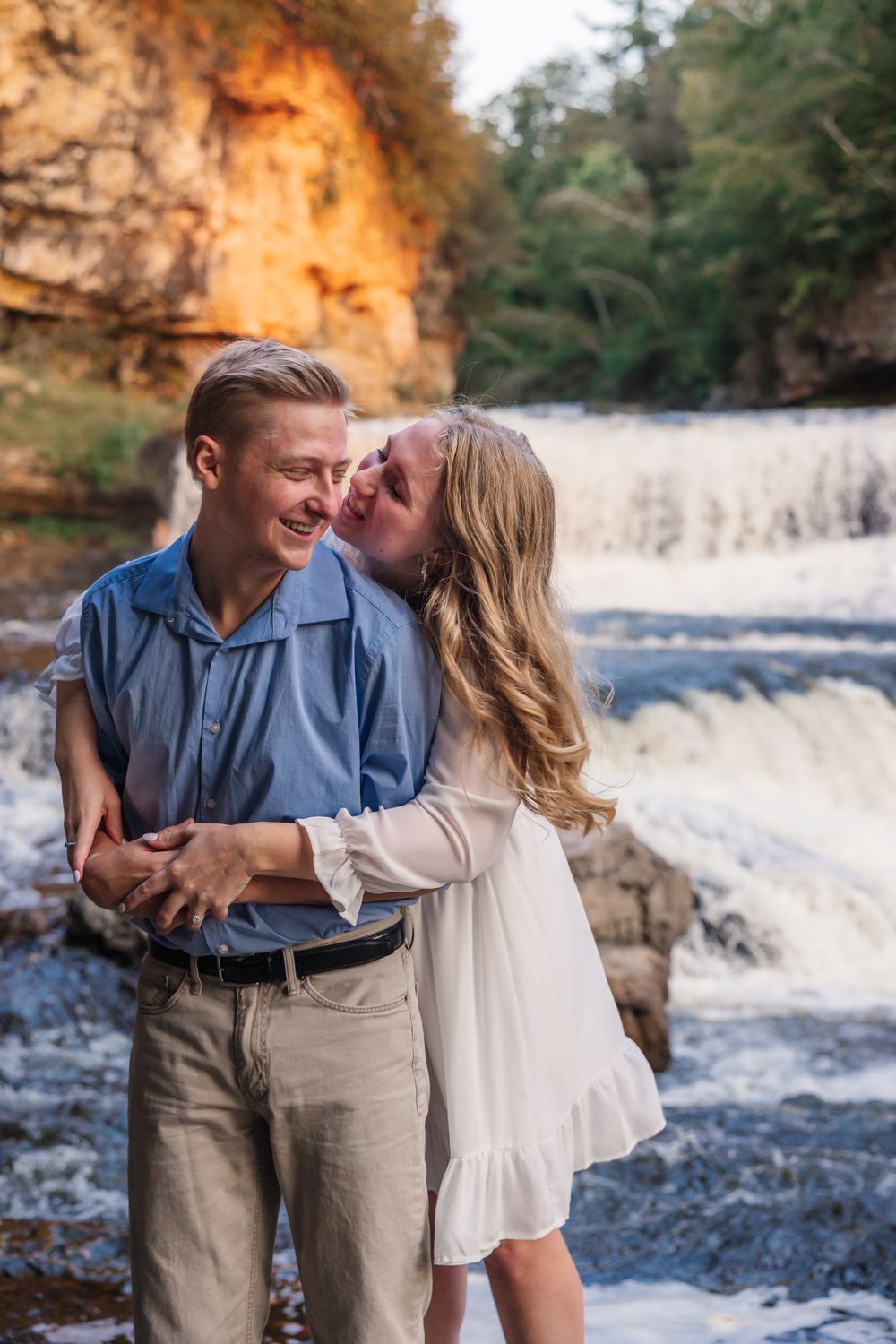 Couple embracing by a scenic waterfall with sunlit cliffs in the background.