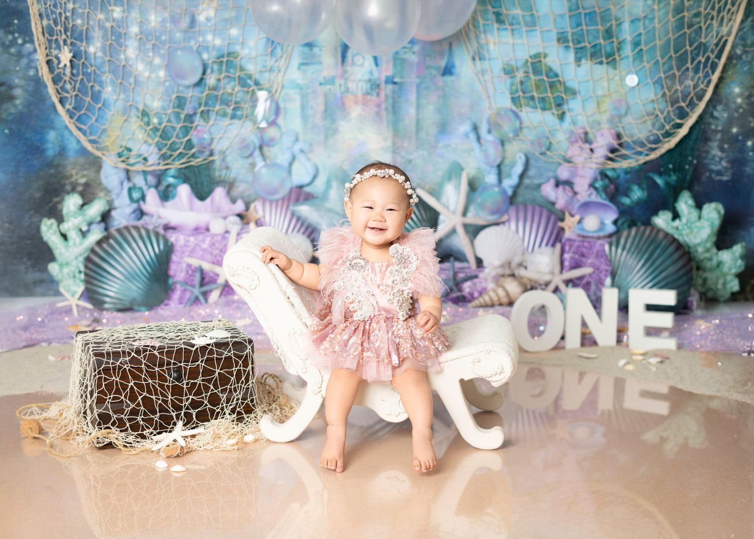 Baby girl smiling, her first birthday cake smash with mermaid theme, pastel balloons, seashell decor, and custom styled backdrop in Covina