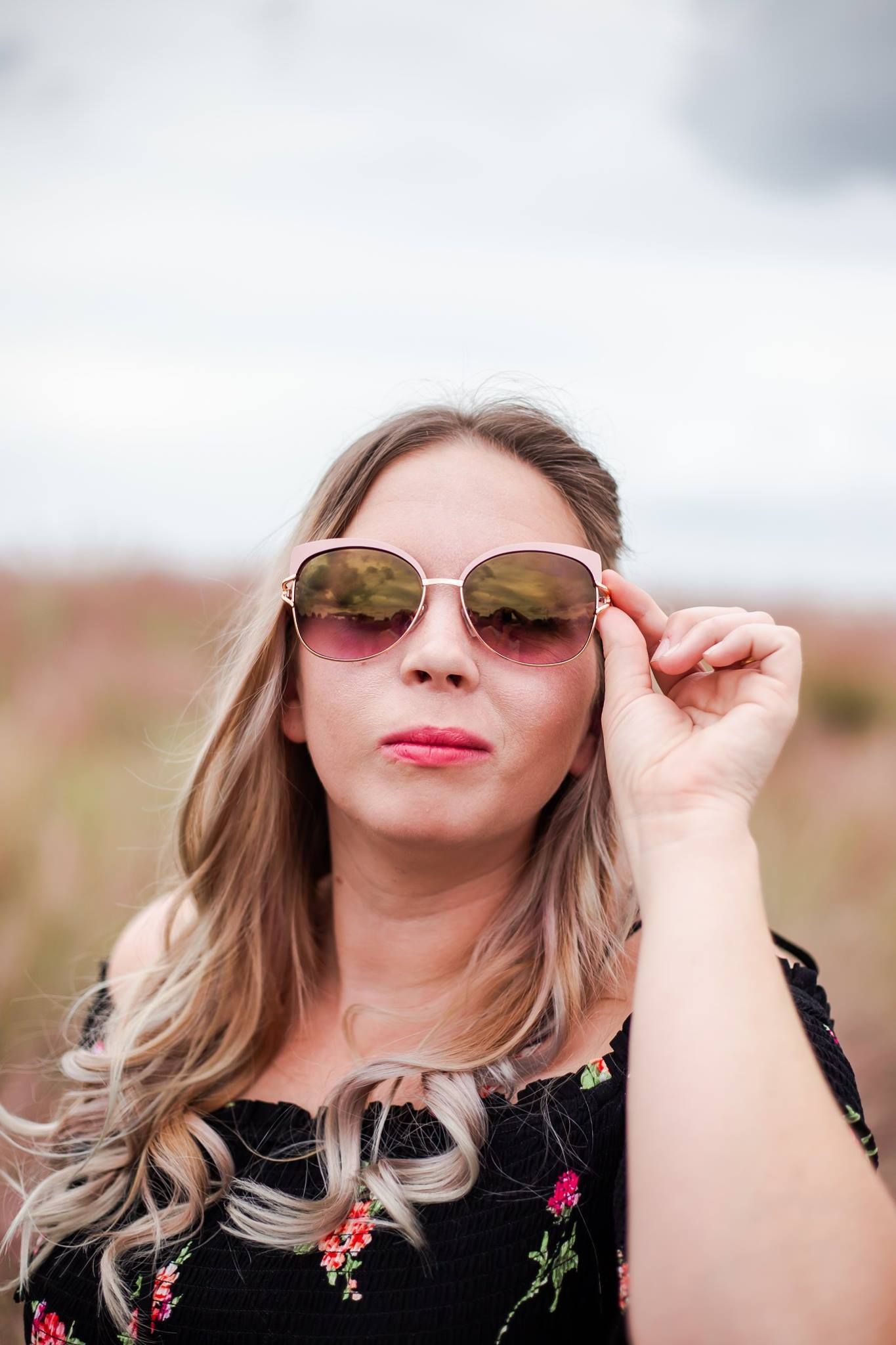 Person in black floral dress adjusts sunglasses while posing in outdoor setting.