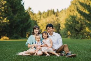 Family relaxing together on green grass in a park during golden hour with evergreen trees in background.