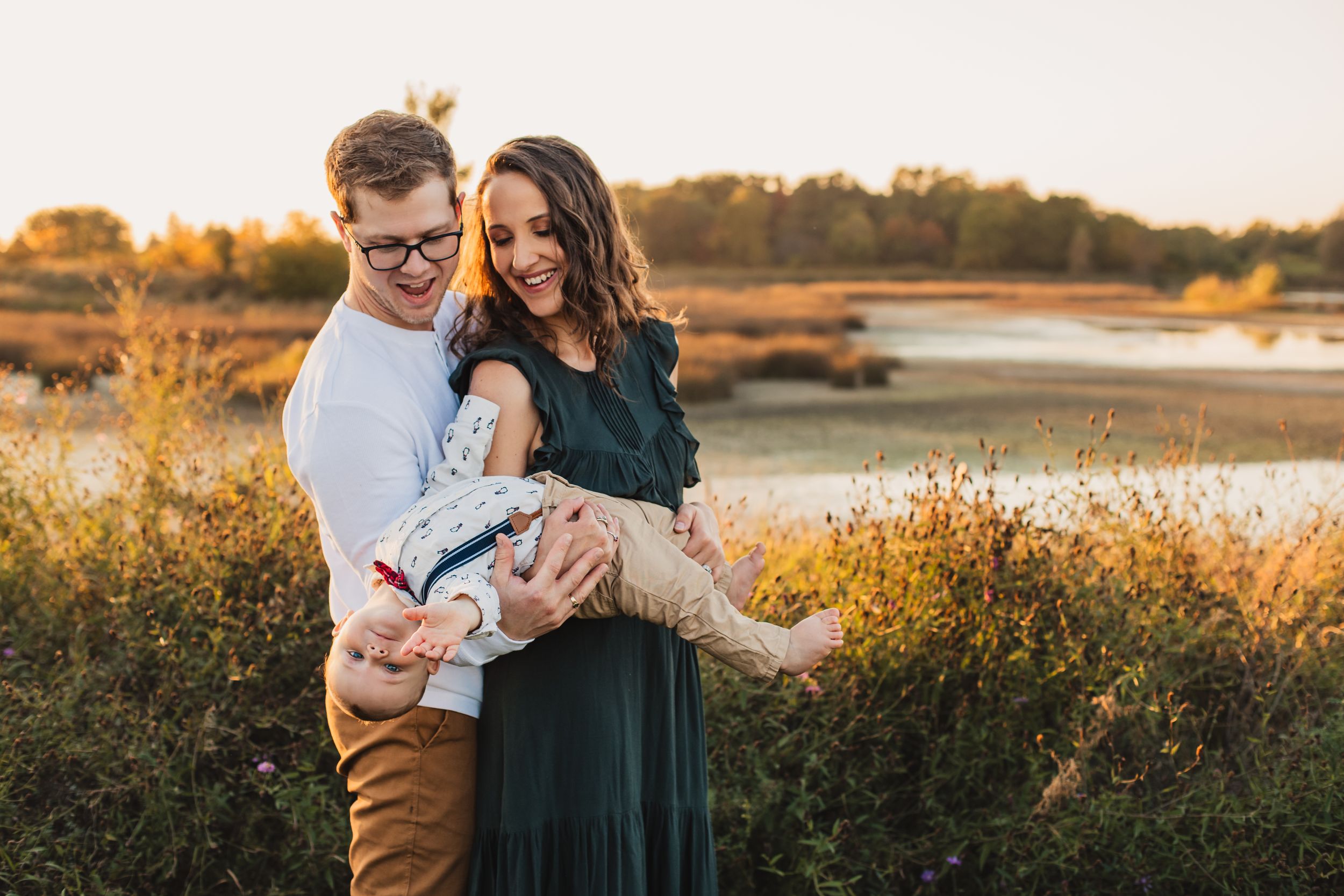 A small family stand in a golden field at sunset beside a lake, creating a fun family outdoor portrait.