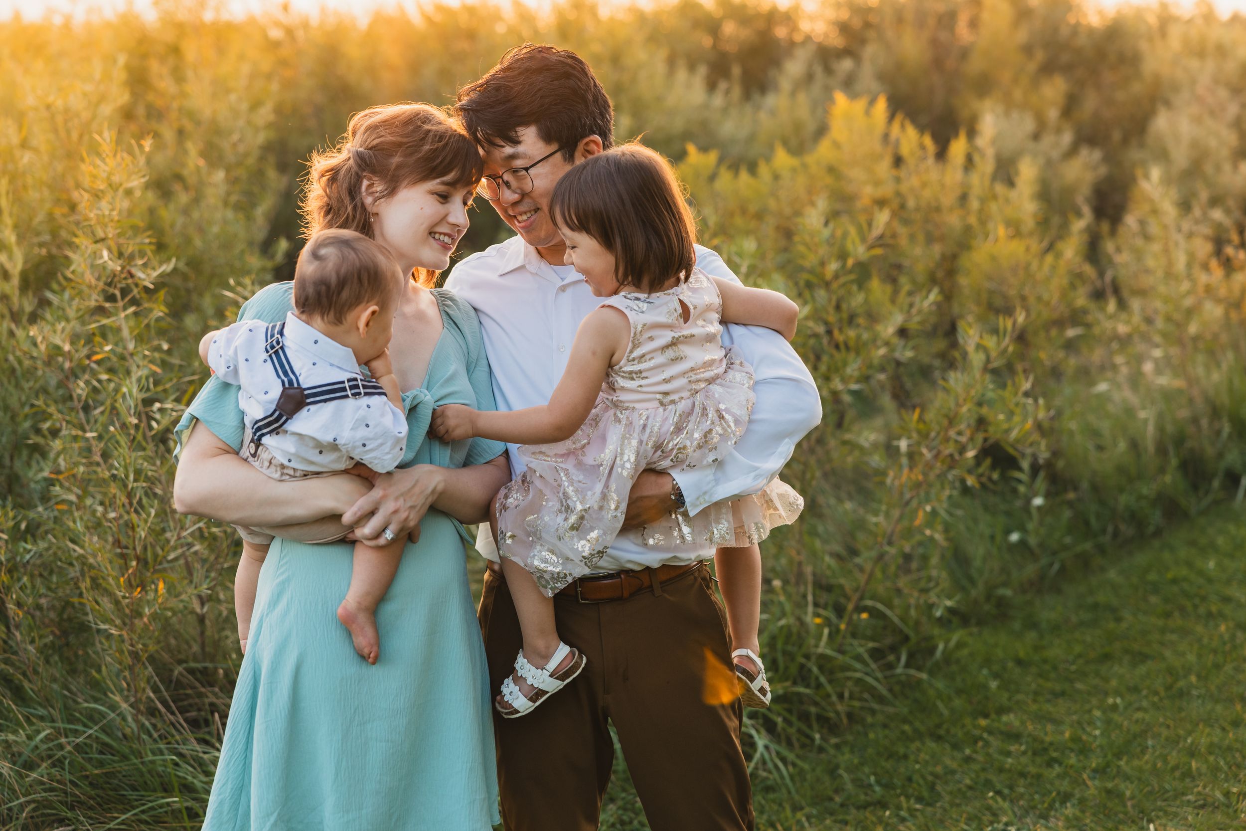 Family wearing coordinated mint and brown outfits embracing during golden hour outdoor photoshoot in tall grass field.