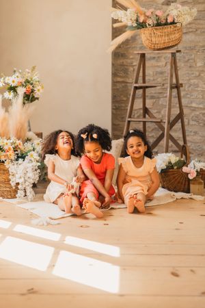 Three children sit together on a wooden floor laughing in a sunlit room decorated with floral arrangements and baskets.