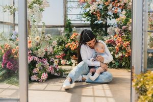 A parent and child sit together in a sunlit greenhouse surrounded by blooming flowers and climbing vines.