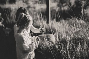 Two girls in white clothing stand in a sunlit meadow of tall grass, captured in black and white to give a feeling of connection.