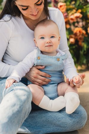 A baby wearing blue overalls and white socks sits on her mother's lap on the greenhouse floor.