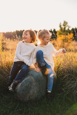 Two children sit together on a large rock in a golden field at sunset, sharing playful laughter and joy.