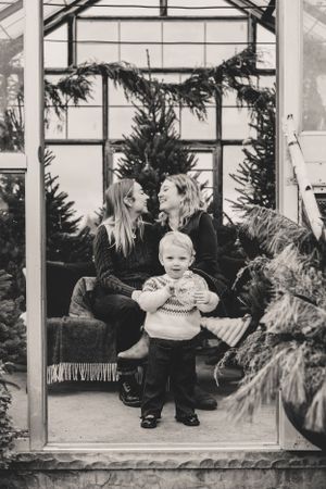 A black and white photo series of a child and their two mothers sitting on a greenhouse bench with evergreen trees in the background.