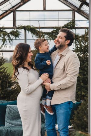 A family shares joyful moments together in a bright holiday greenhouse setting with evergreen trees in the background.