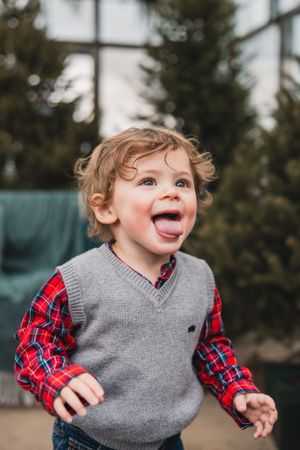Child wearing gray vest and red plaid shirt laughs joyfully while playing in the greenhouse with evergreen trees in background.