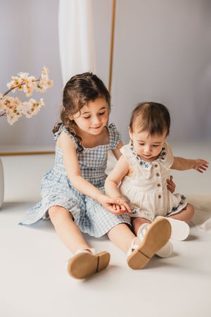 Two children in gingham and white dresses play together on a light-colored floor with floral decorations.