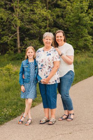 Three generations of women in casual summer outfits walk together on a gravel path surrounded by lush green foliage.