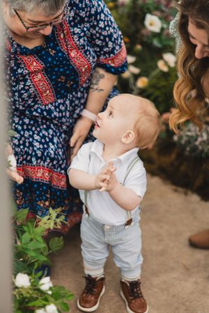 A toddler in white outfit and suspenders stands in a greenhouse while looking upward towards his grandmother.