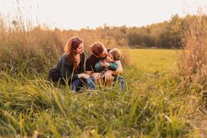 Two people and their toddler sit together in a golden field during sunset while sharing a snuggle.