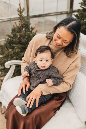 A parent in a beige sweater holds a baby wearing a gray knit sweater while sitting on a white bench in a greenhouse.