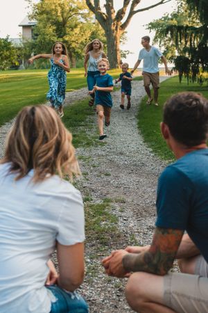 Parents kneel and watch their children play and run in a yard setting with trees in the background.