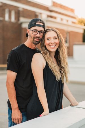 A couple in casual black outfits lean against a white ledge during a sunset photoshoot with warm lighting.