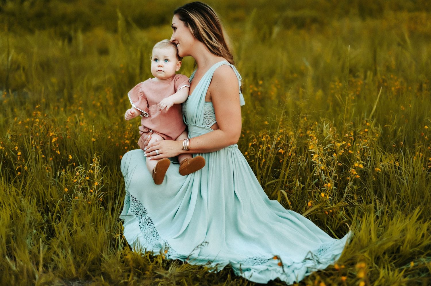 A woman in a flowing mint dress sits in a golden meadow with a baby wearing pink during sunset.
