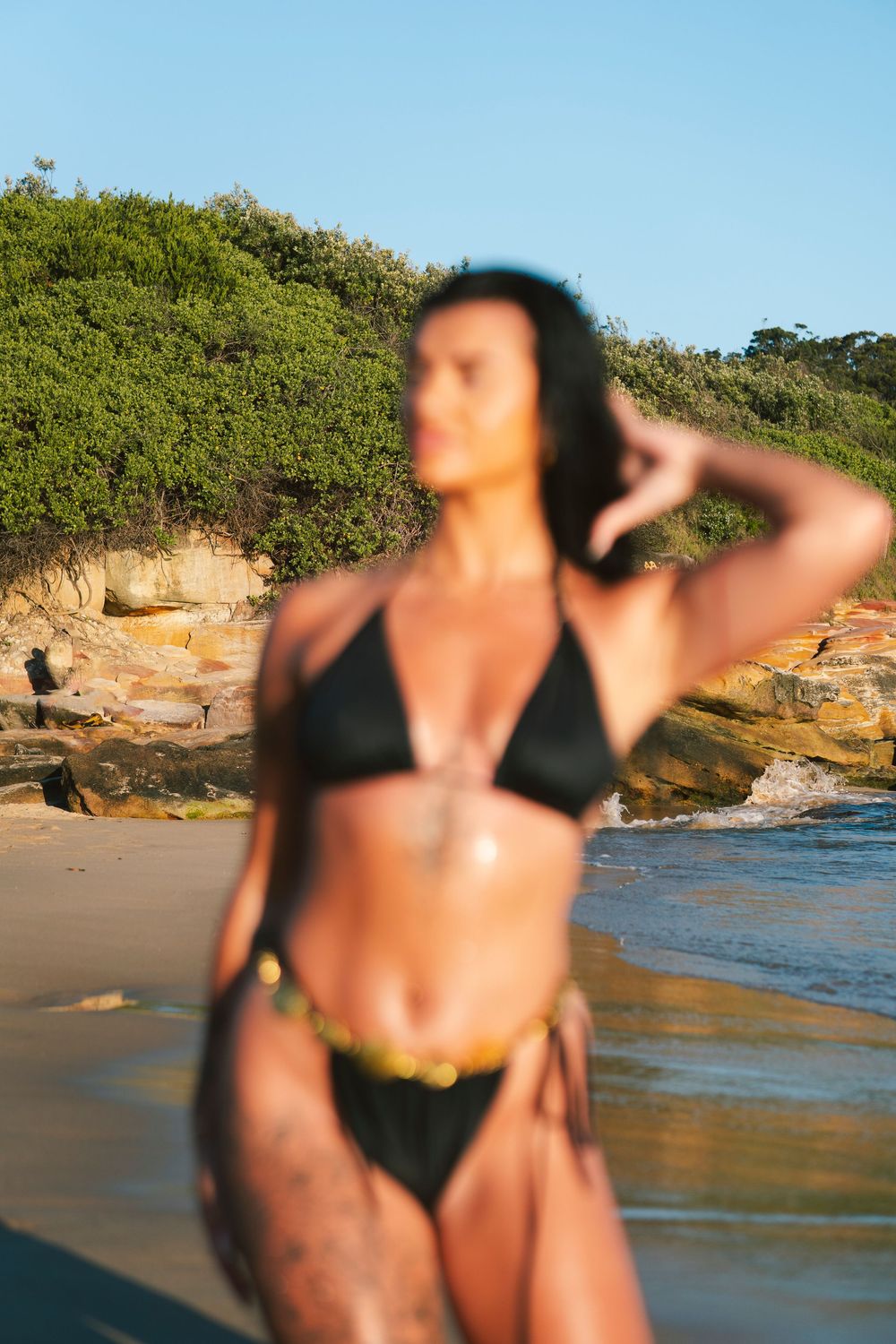 Woman in black bikini posing on sandy beach with rocky cliffs and green vegetation in background.