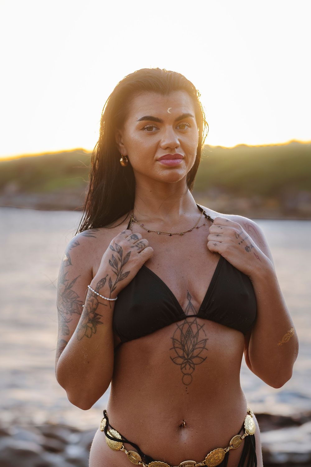 Tattooed woman in black bikini with gold belt poses confidently at golden hour beach, looking at camera.