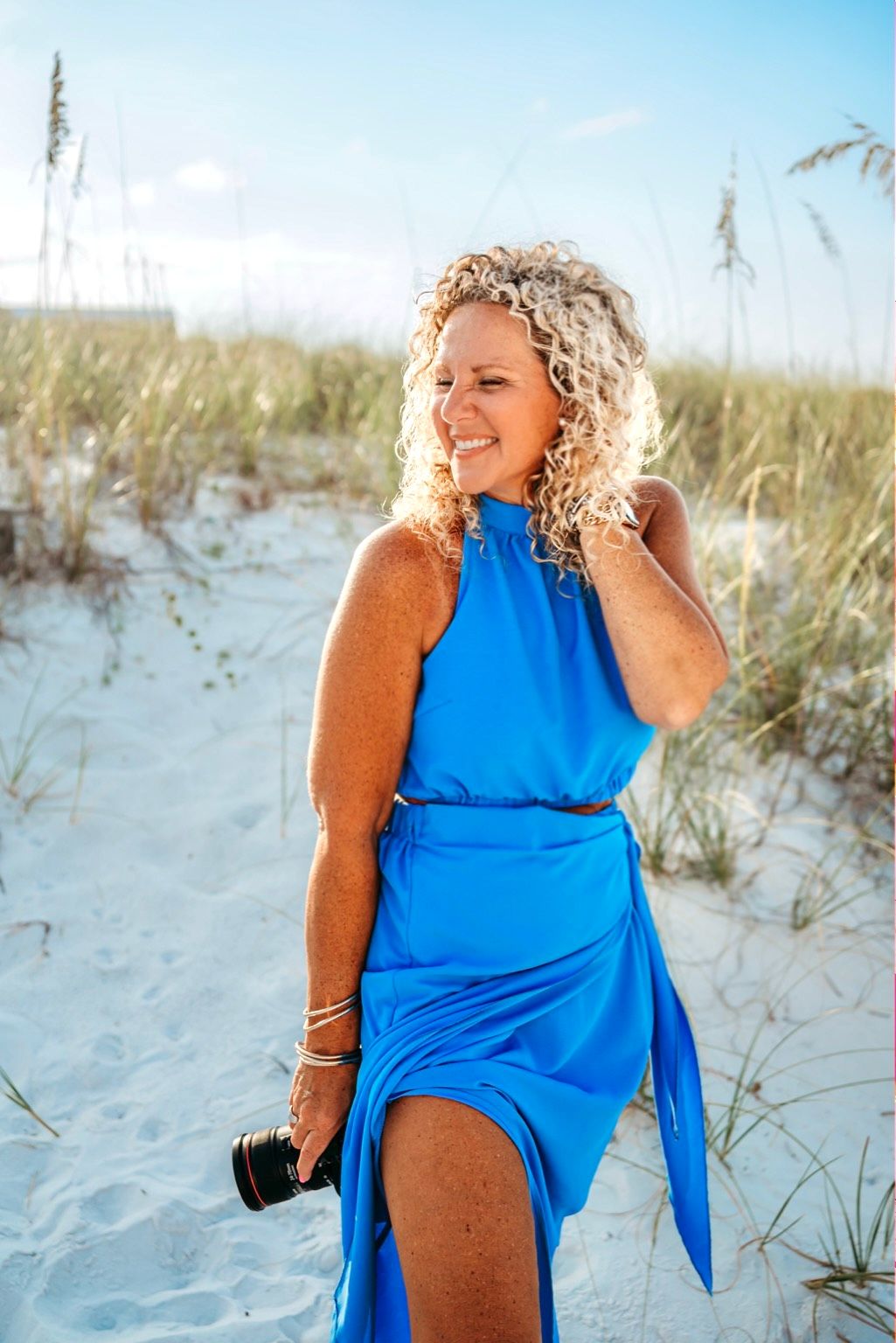 Woman in bright blue swimsuit cover up poses on sandy beach with sea grass and dunes in background.