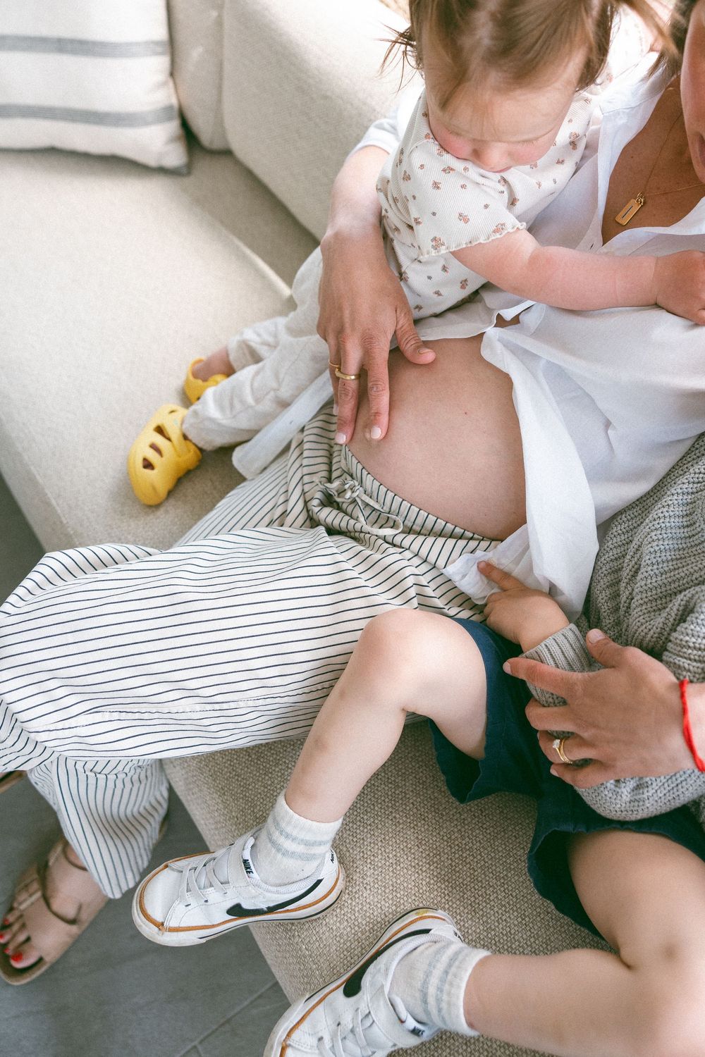 A toddler touches a pregnant woman's bare belly while sitting on a couch with another person nearby.