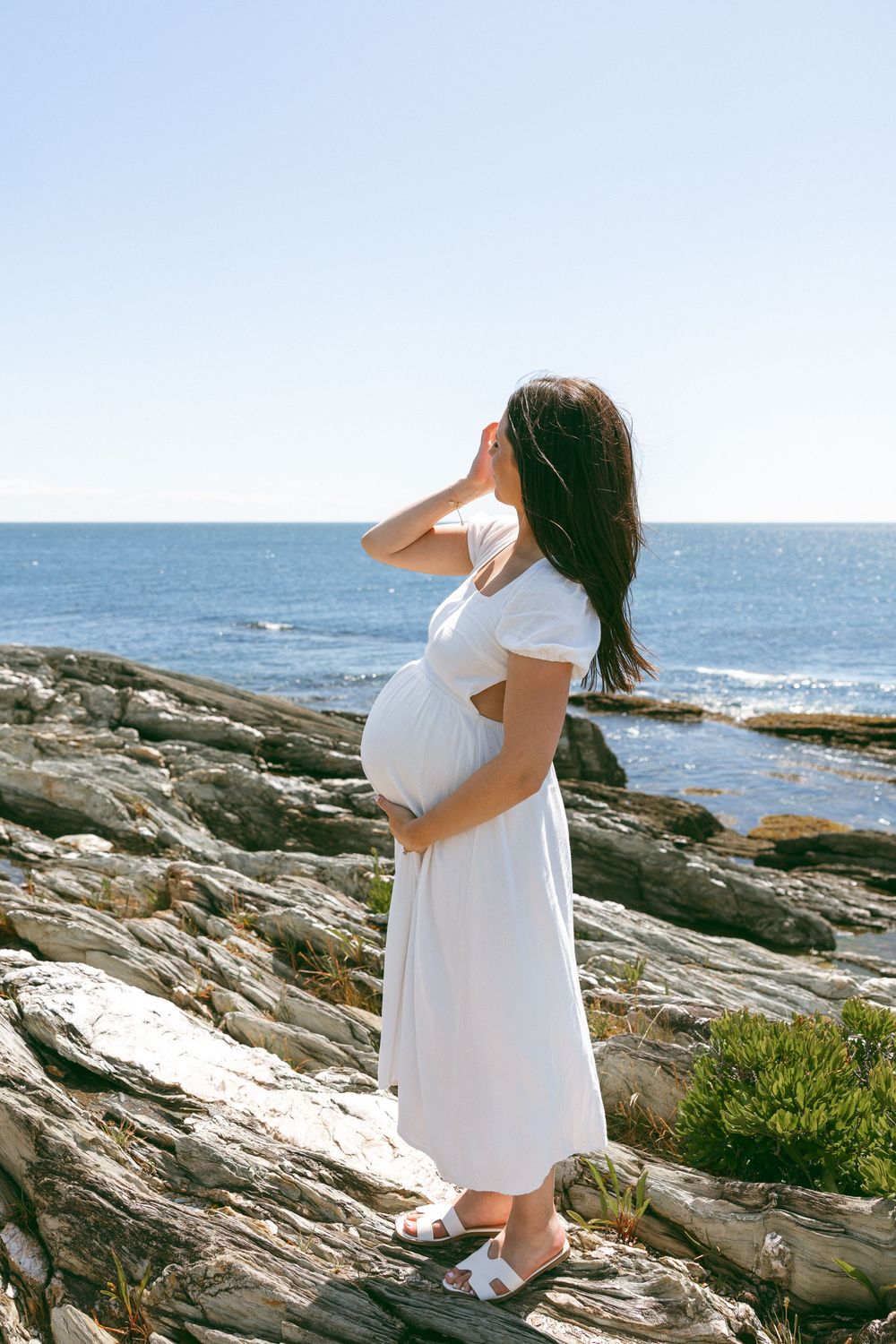 Person in white dress poses on rocky coastal cliffs overlooking the ocean.